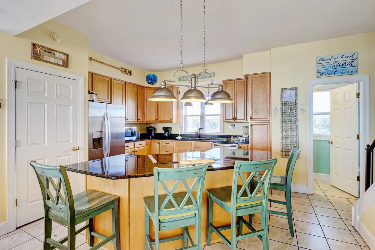 A bright, open kitchen scene featuring a central kitchen island with bar stools, modern cabinetry, and a range of stainless steel appliances. The space is arranged for both cooking and casual dining, inviting for family gatherings or prepping meals.