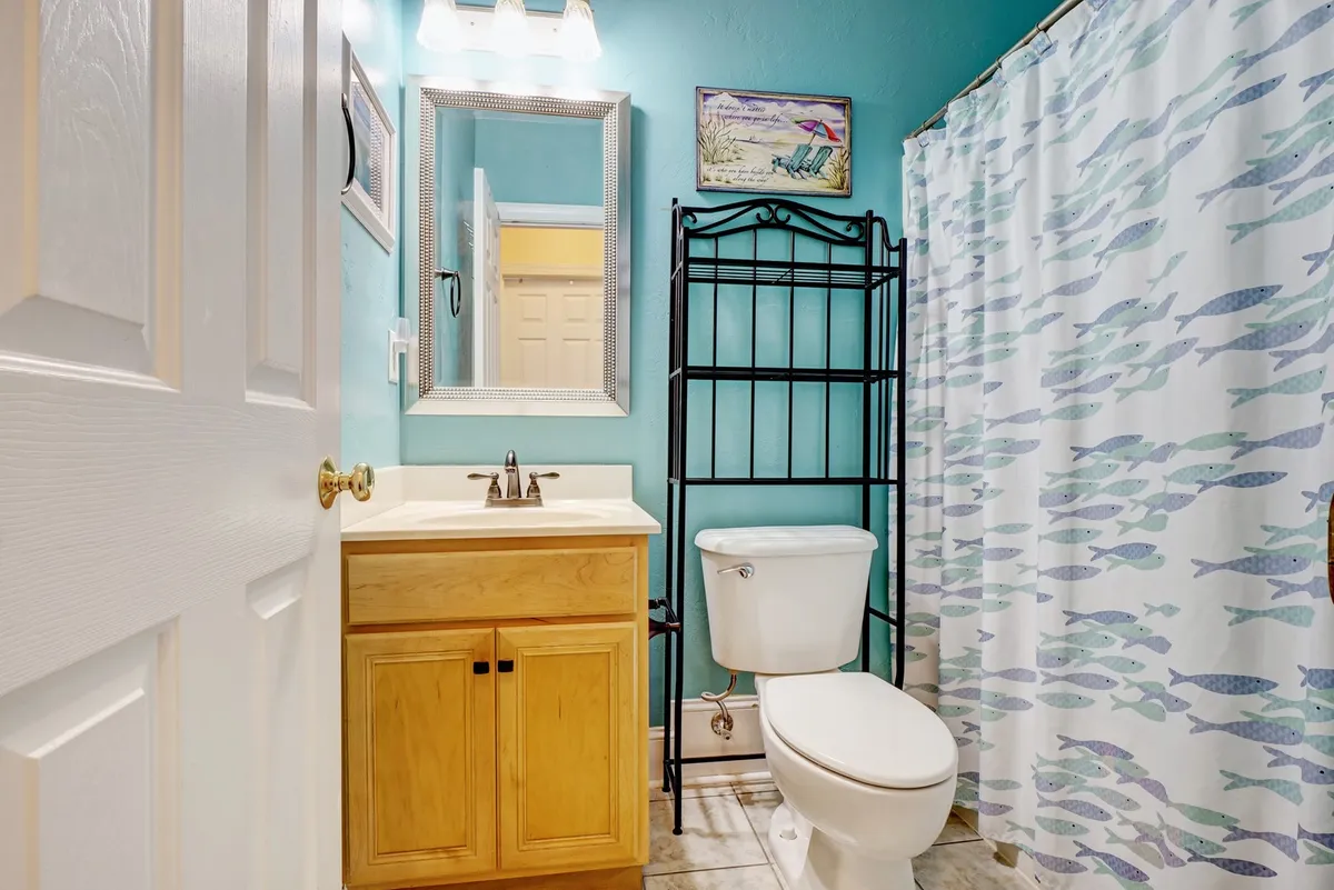 A bathroom with a cheerful, beach-inspired design, featuring a fish-patterned shower curtain, wooden vanity, and a mirror with a decorative frame. The space feels fresh and welcoming—ideal for prepping for a new day or winding down after adventures.