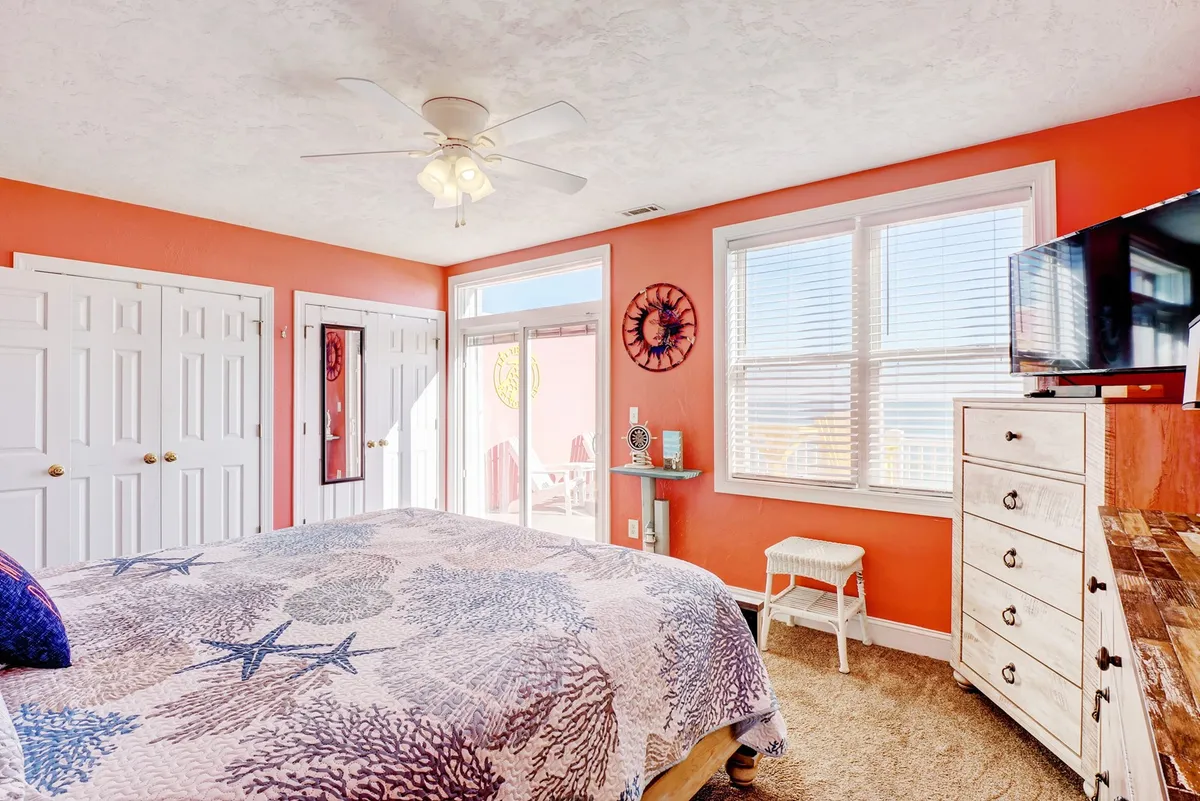 A serene bedroom decorated with coral-themed bedding, large windows providing ample natural light, and tasteful decorative wall art. The room exudes a calm and peaceful mood, perfect for relaxing after a day at the beach.