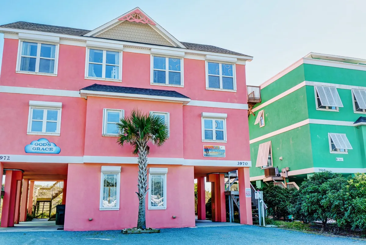 A vibrant pink building facade stands out under clear skies, flanked by a tall palm tree. The sign "GOD'S GRACE" adds a welcoming touch, inviting guests to the property. The scene hints at a cheerful, relaxing vacation environment, ideal for family gatherings and outdoor relaxation.