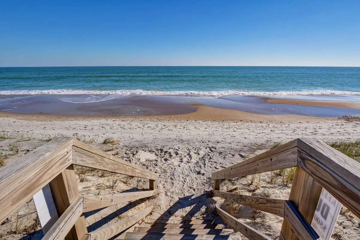 This image shows broad wooden stairs descending directly onto sandy beach, with the ocean stretching out under a wide, open sky. The space feels serene and inviting, perfectly capturing the sense of direct, private access from the residence to the shoreline. You can see the transition from the deck area to the beach, emphasizing the ease of stepping out and enjoying the seaside.