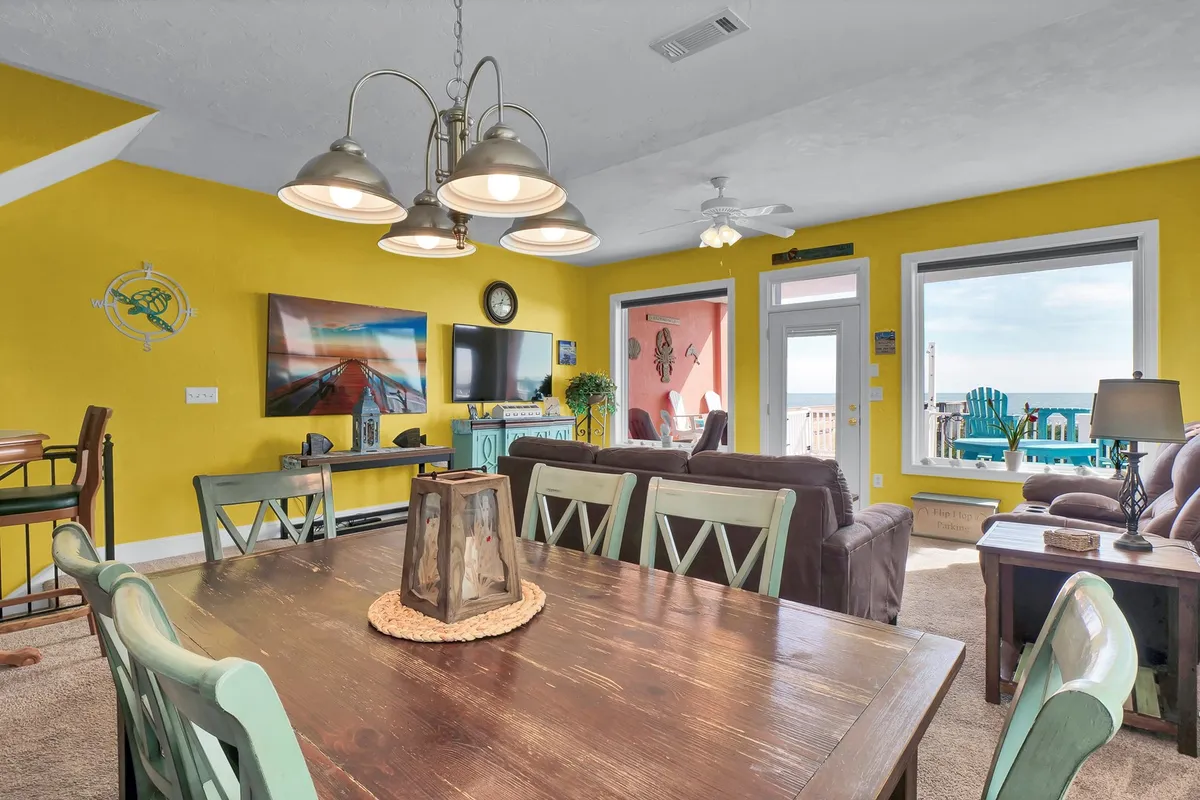 A casual, welcoming open-concept living and dining area featuring a large dining table, vibrant walls, and expansive windows that offer direct views of the ocean. The entertainment center with a flat-screen TV and comfortable seating encourages relaxation and family gatherings.