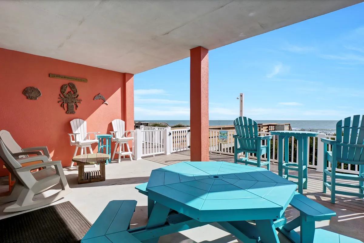 A cheerful outdoor area with a turquoise table and white Adirondack chairs overlooks the ocean. The coral wall adds a playful pop of color, and the view suggests a space for relaxing after a day of beach play, perhaps while drying off pets or enjoying the sea breeze.