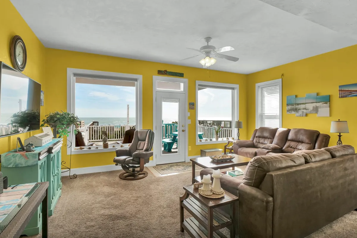 A bright living area with comfortable brown sofas, vibrant yellow walls, and large windows framing stunning ocean views. The setting is ideal for relaxing, socializing, or enjoying a board game with friends and family.