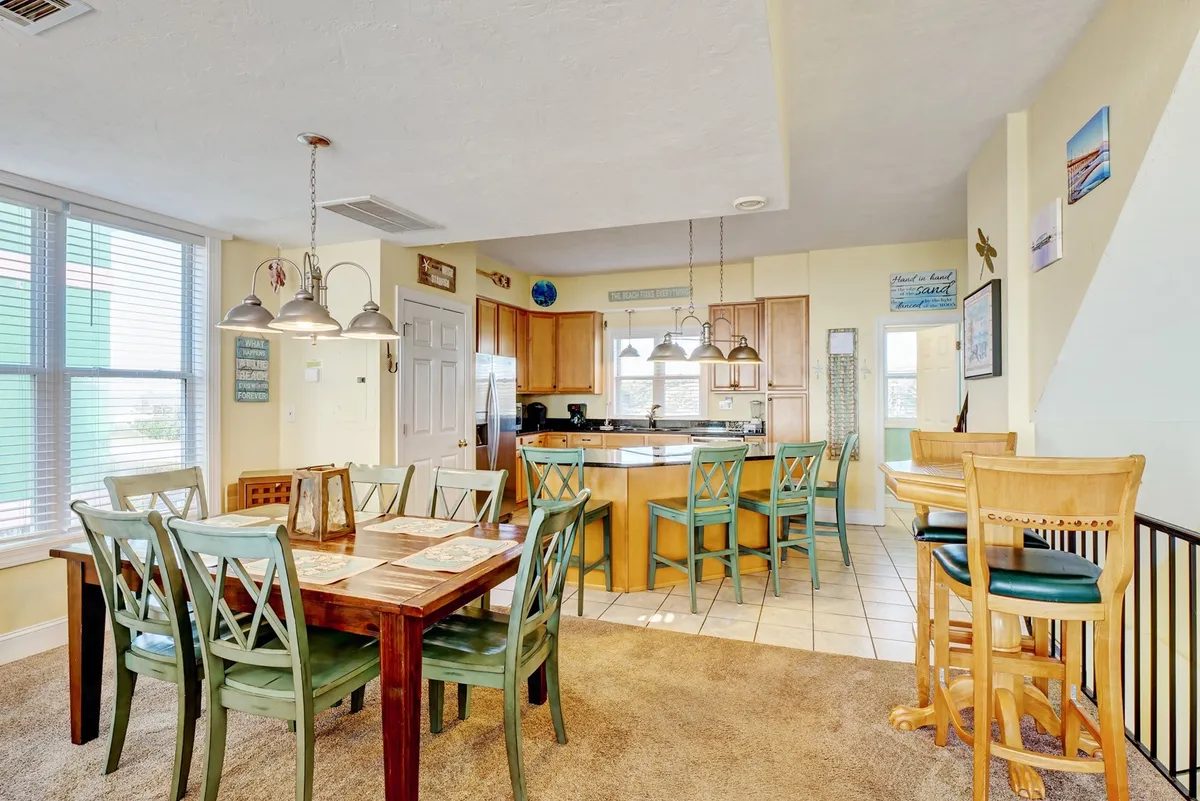 A cheerful interior with a dining table and bar stools adjacent to a kitchen island, set against large windows that bring in natural light. The scene suggests a versatile space suited to social meals and casual mornings.