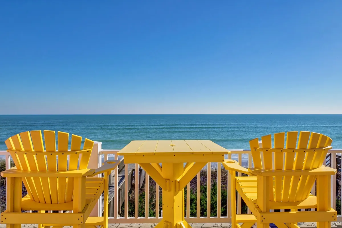 The image showcases the exterior deck of a single-family beachfront house on North Topsail Beach, featuring inviting yellow chairs and a table overlooking sweeping ocean views. The tranquil scene suggests an ideal spot for sunrise breakfasts, sunset cocktails, and peaceful moments with loved ones.