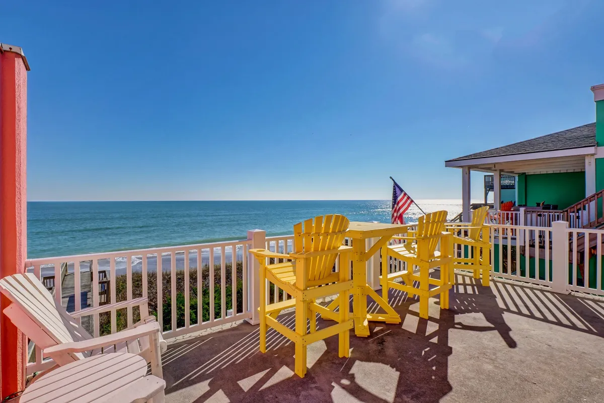The image captures a cheerful and tranquil deck area with yellow outdoor seating facing the ocean. The colorful facade of the house, an American flag waving gently, and expansive water views evoke a classic coastal escape. The scene feels welcoming and perfect for social gatherings or relaxing moments watching the sunset.
