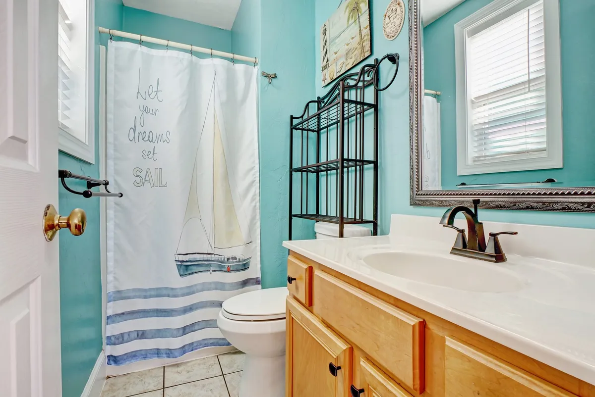 This image shows a bathroom with a cheerful sailboat-patterned shower curtain, a sink with a cabinet, a mirror, and decorative wall art. The setting is bright and uplifting, evoking the sense of fresh, beachside living and cleanliness.