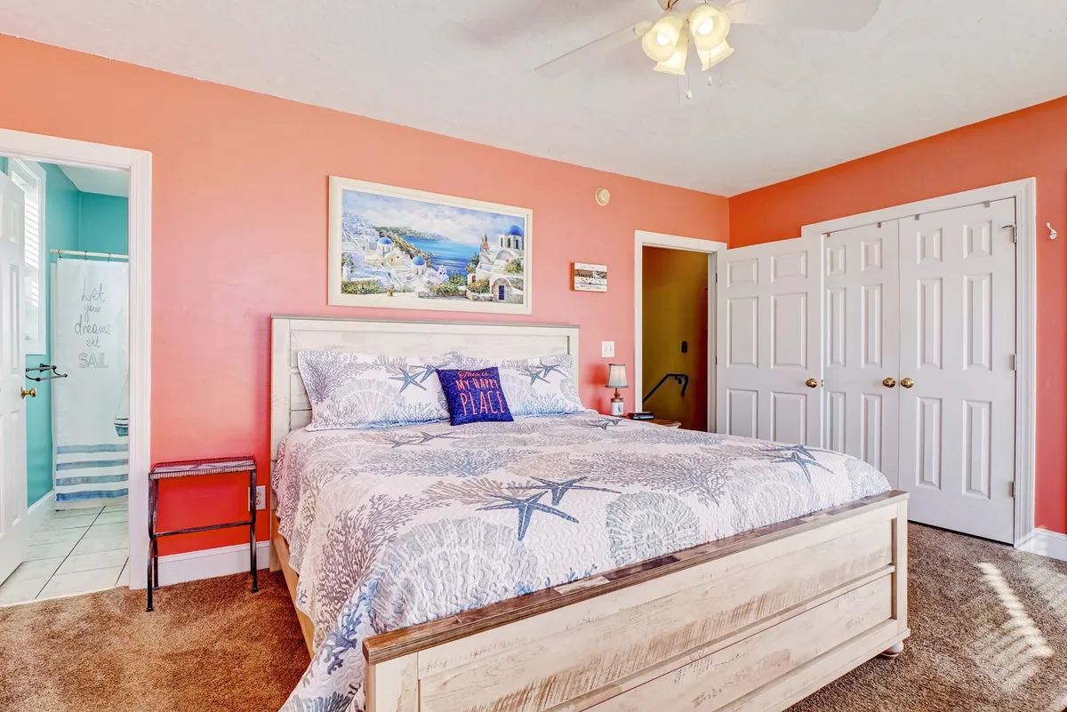 A cozy bedroom featuring a bed with a starfish-patterned blanket, colorful wall art, a comfortable chair, and a nightstand with a lamp. The room feels bright, cheerful, and inviting, offering a welcoming space for relaxation and reading.