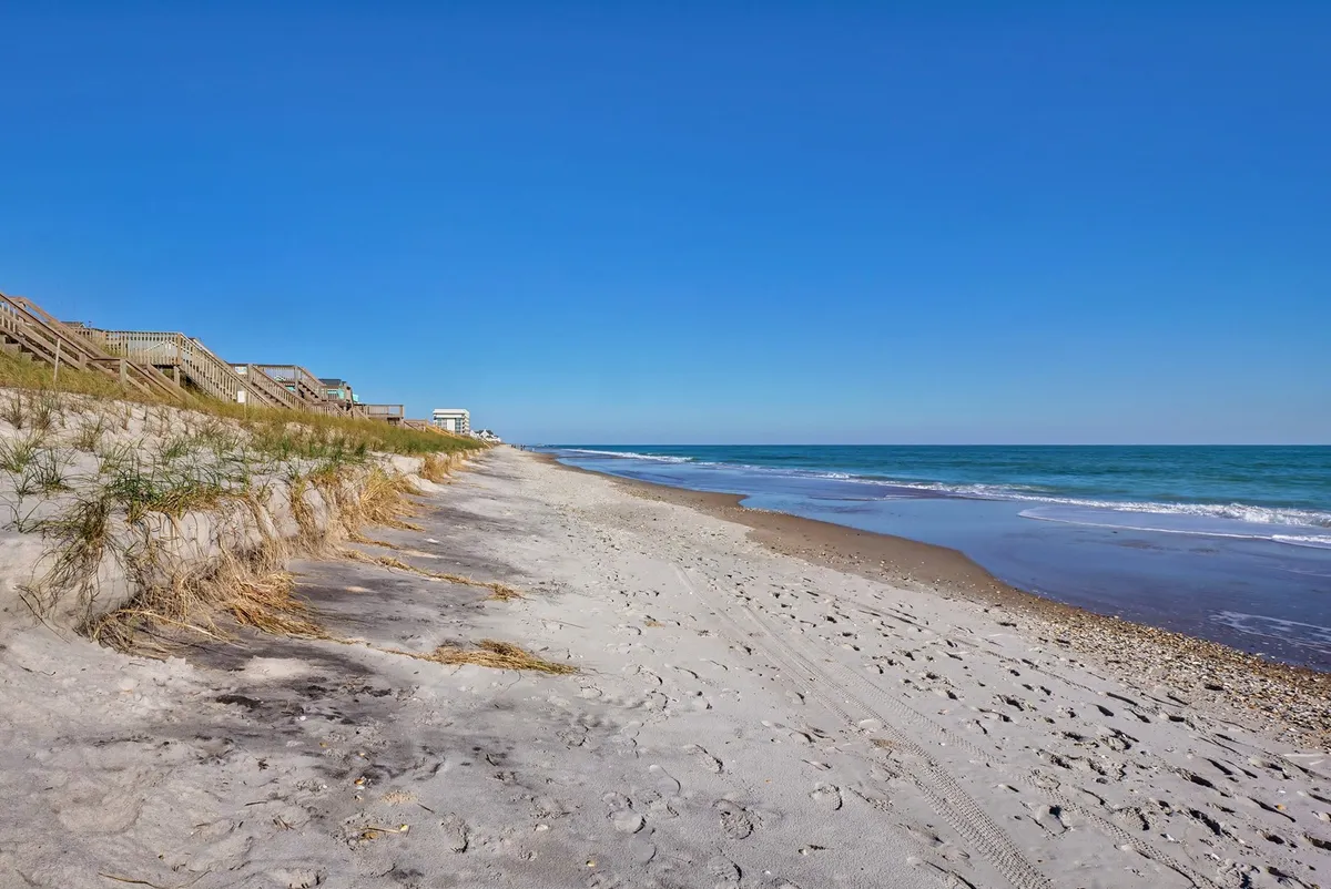 A wide view of North Topsail's sandy beach dotted with dune grass, with gentle ocean waves and a boardwalk leading out toward the water. People can be seen enjoying the natural beauty—walking, sunbathing, and taking in the relaxing atmosphere. The scene is open and expansive, perfect for families and groups to spread out and enjoy various beach activities.